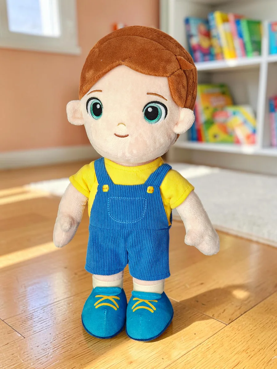 Levi, la poupée garçon Playnitive, debout sur le parquet d’une chambre d’enfant : cheveux châtains, t-shirt jaune et salopette en velours côtelé bleu